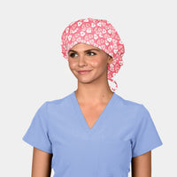 Person wearing a light blue scrub top and a pink scrub cap with heart pattern on a plain background
