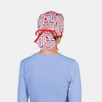 woman wearing a blue scrub top and a ponytail scrub hat that is pink with words and red hearts on it in a breast cancer awawreness theme