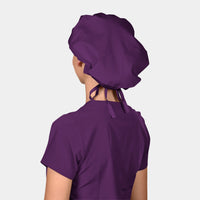 A person wearing a deep purple scrub hat, matching the Eggplant shade, from the back and a matching scrub top on a light grey background.