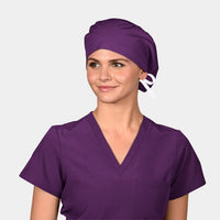 A person wearing a solid purple scrub hat with a bow on the top, matching with a purple v-neck scrub set.