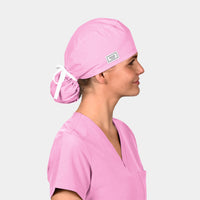 A person wearing a pink ponytail scrub hat tied at the back, matching with a pink scrub top on a light grey background.