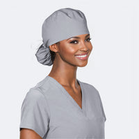 A person wearing a slate grey scrub hat with a ponytail and a grey scrub top on a light grey background.