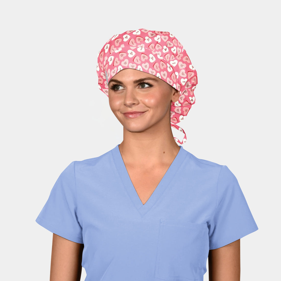 Person wearing a light blue scrub top and a pink scrub cap with heart pattern on a plain background