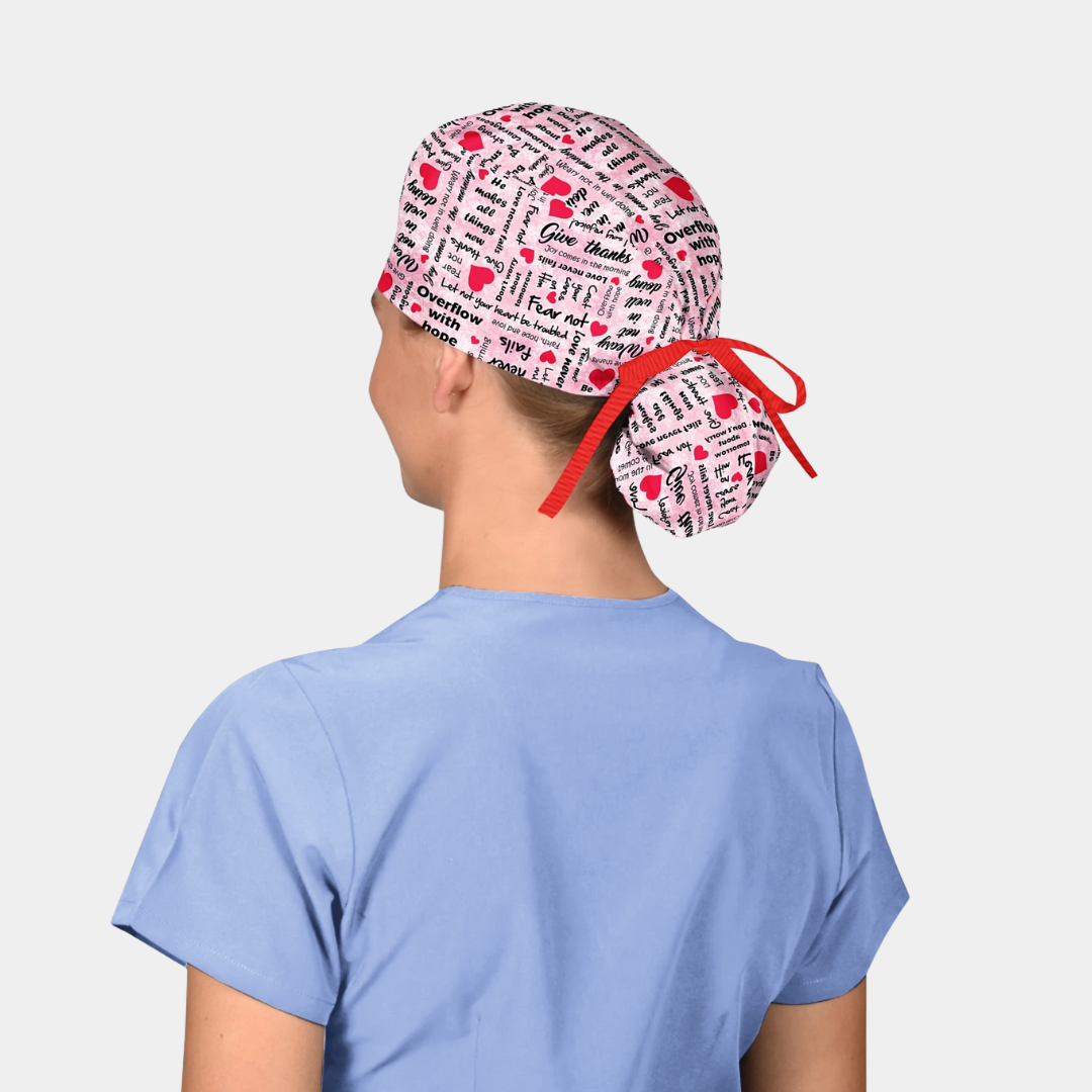 person wearing a ponytail scrub hat with a breast cancer awareness theme and is pink with words and red hearts on it