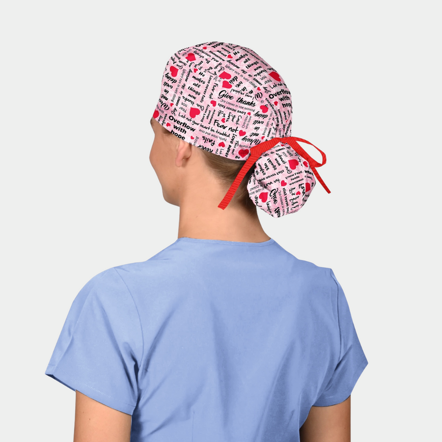 person wearing a ponytail scrub hat with a breast cancer awareness theme and is pink with words and red hearts on it