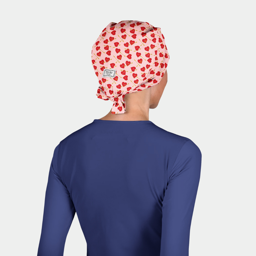 Person wearing a blue scrub top and a red heart patterned scrub cap on a light gray background