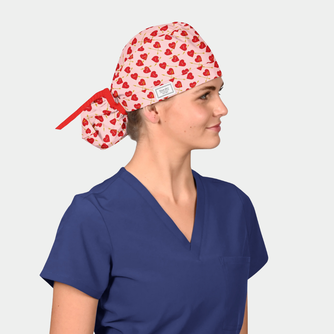 Person wearing a blue scrub top and a patterned scrub cap with red hearts on a light gray background