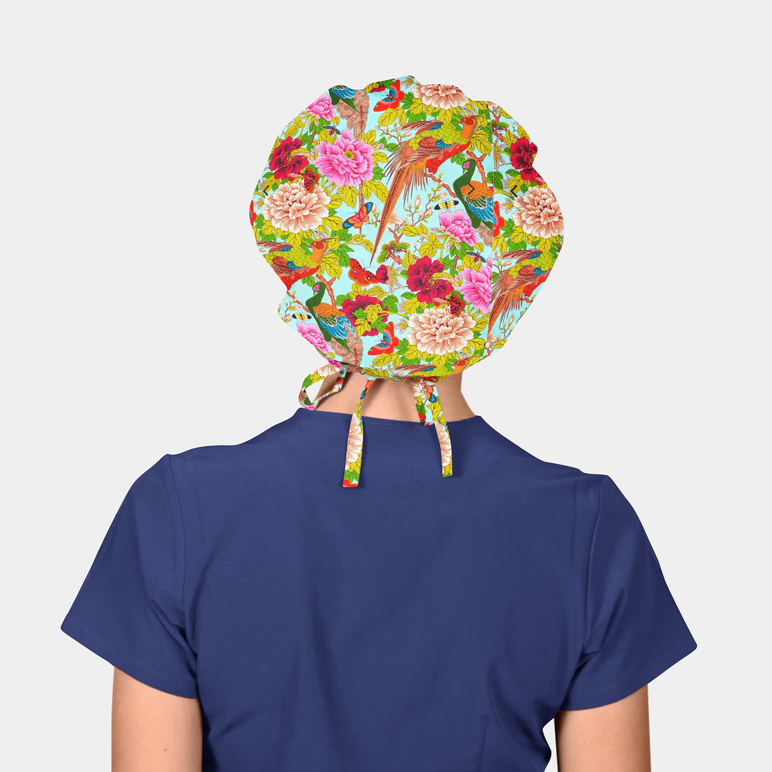 person wearing navy blue scrubs and a colorful bouffant hat with birds and flowers and tress on a light blue background
