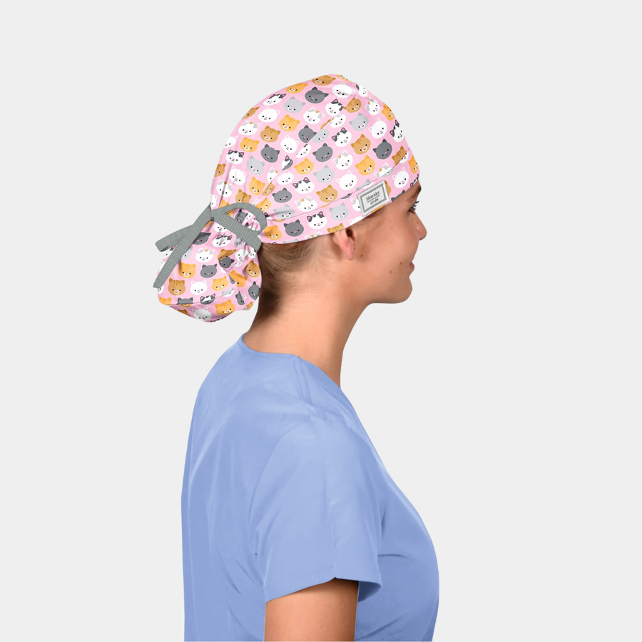 woman wearing a light blue scrub top and a ponytail scrub hat that is pink with cats on it