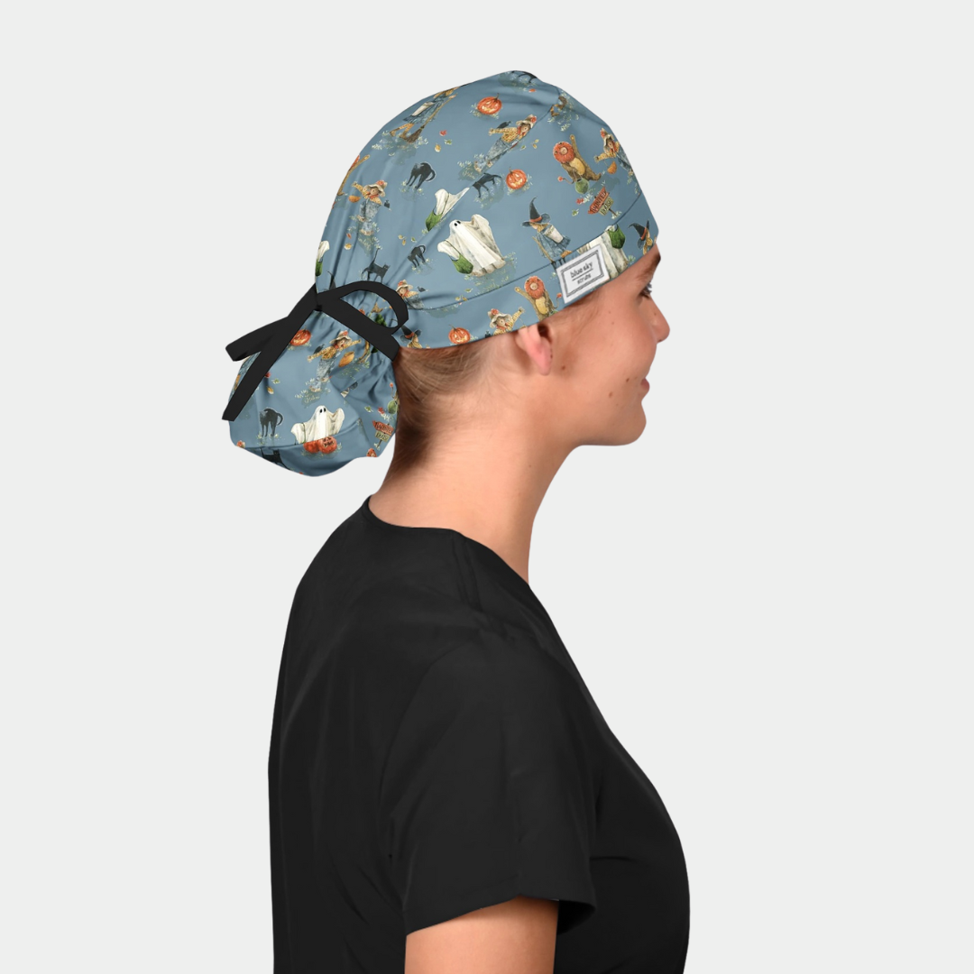 woman wearing a black scrub top and a ponytail style halloween scrub hat that is blue with ghosts, pumpkins and cats 