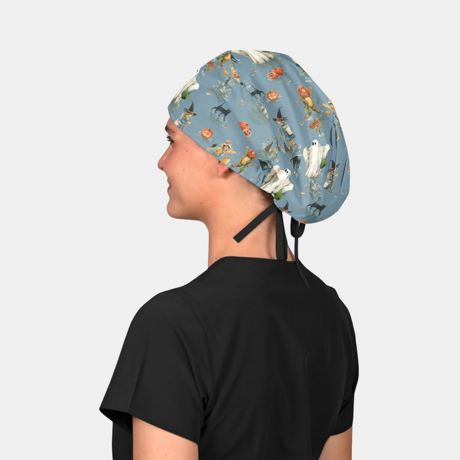 woman wearing a black scrub top and a surgical scrub cap that is halloween themed with cats, pumpkins and ghosts on a blue background