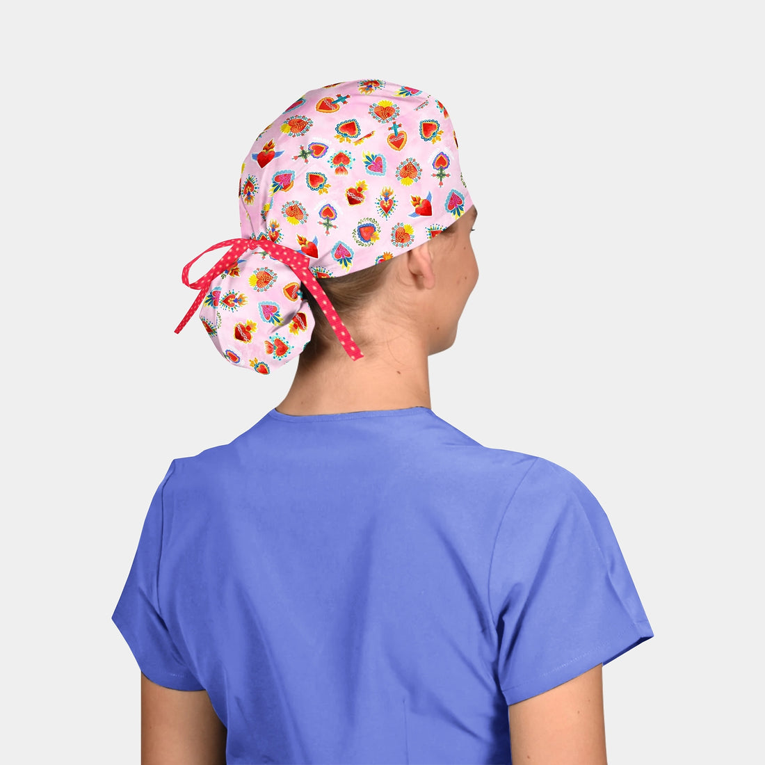 A surgical hat with a light pink background featuring a pattern of colorful hearts, crosses, and crowns. The hat has a red and white bow tie at the back.