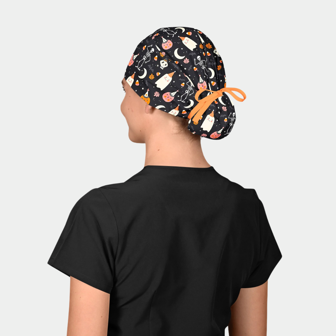 woman wearing a black scrub top and a halloween scrub cap with ghosts, skeletons and pumpkins on a black backgound.