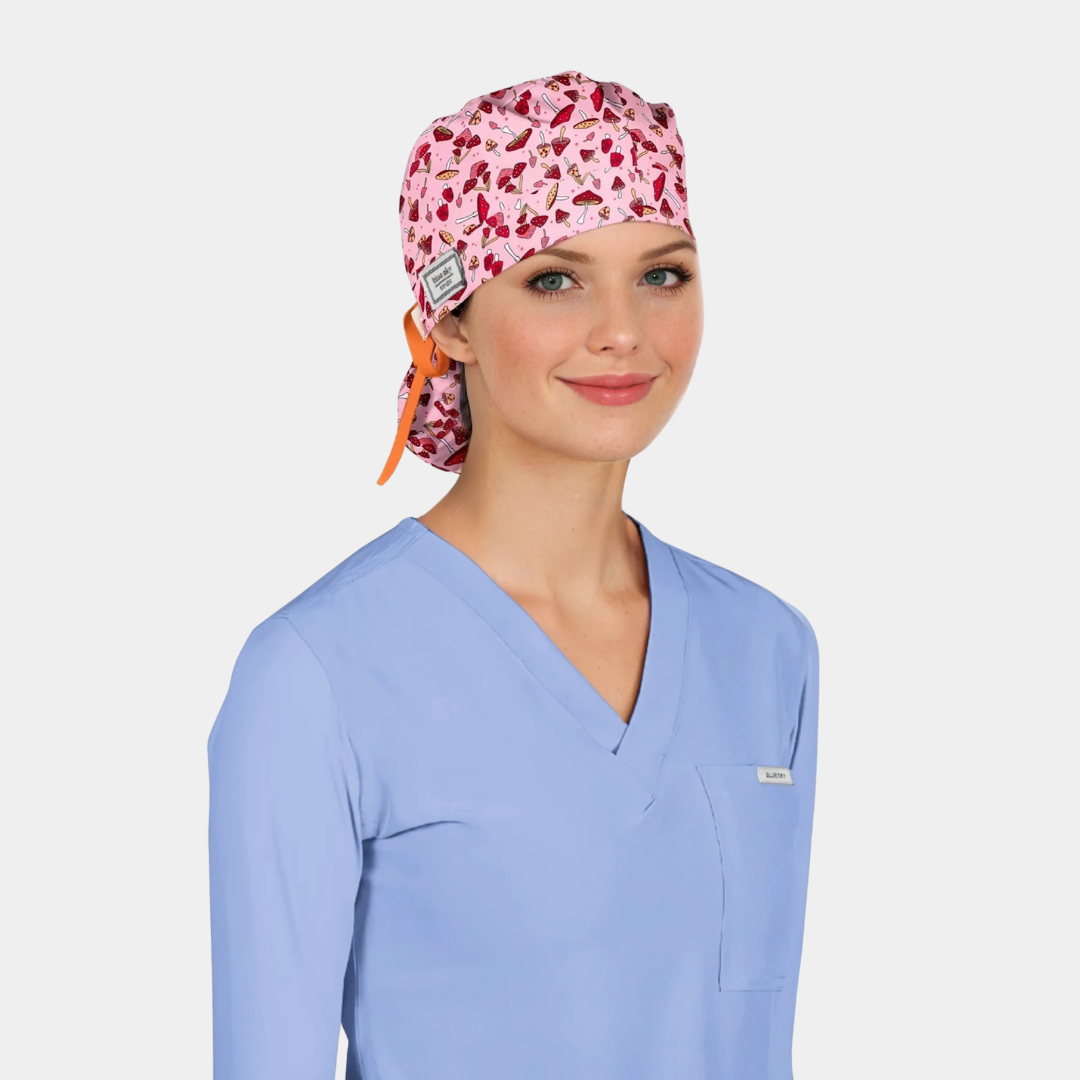Woman wearing a light blue scrub top and a pink scrub cap with red mushrooms on it on a white background